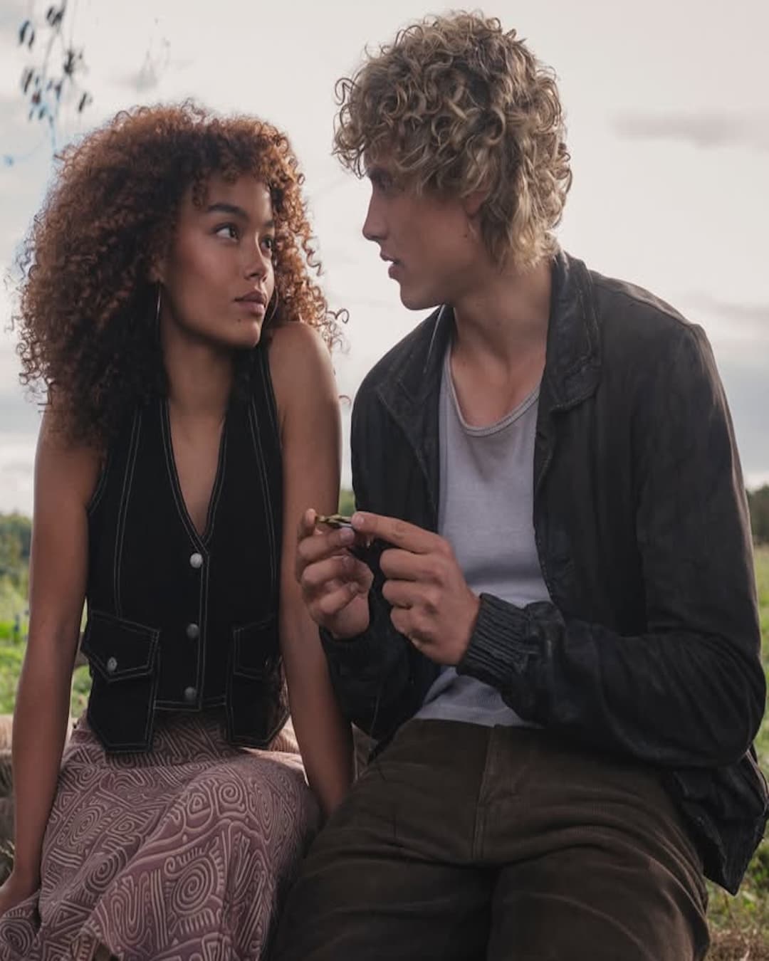 A young woman with curly hair and a patterned skirt sits outdoors next to a young man with blonde curls and a dark jacket. They share an intent look as he holds a small object, evoking the heartfelt moments seen in trending BookTok YA adaptations.
