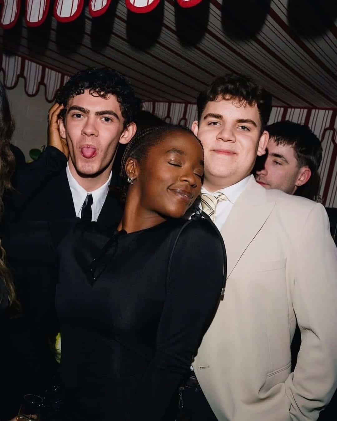 Three young people pose at a party. The woman in front smiles with her eyes closed, leaning on a man in a light suit who is smiling. Another man behind her makes a playful face—like they're celebrating the next big 2026 YA adaptations on BookTok.