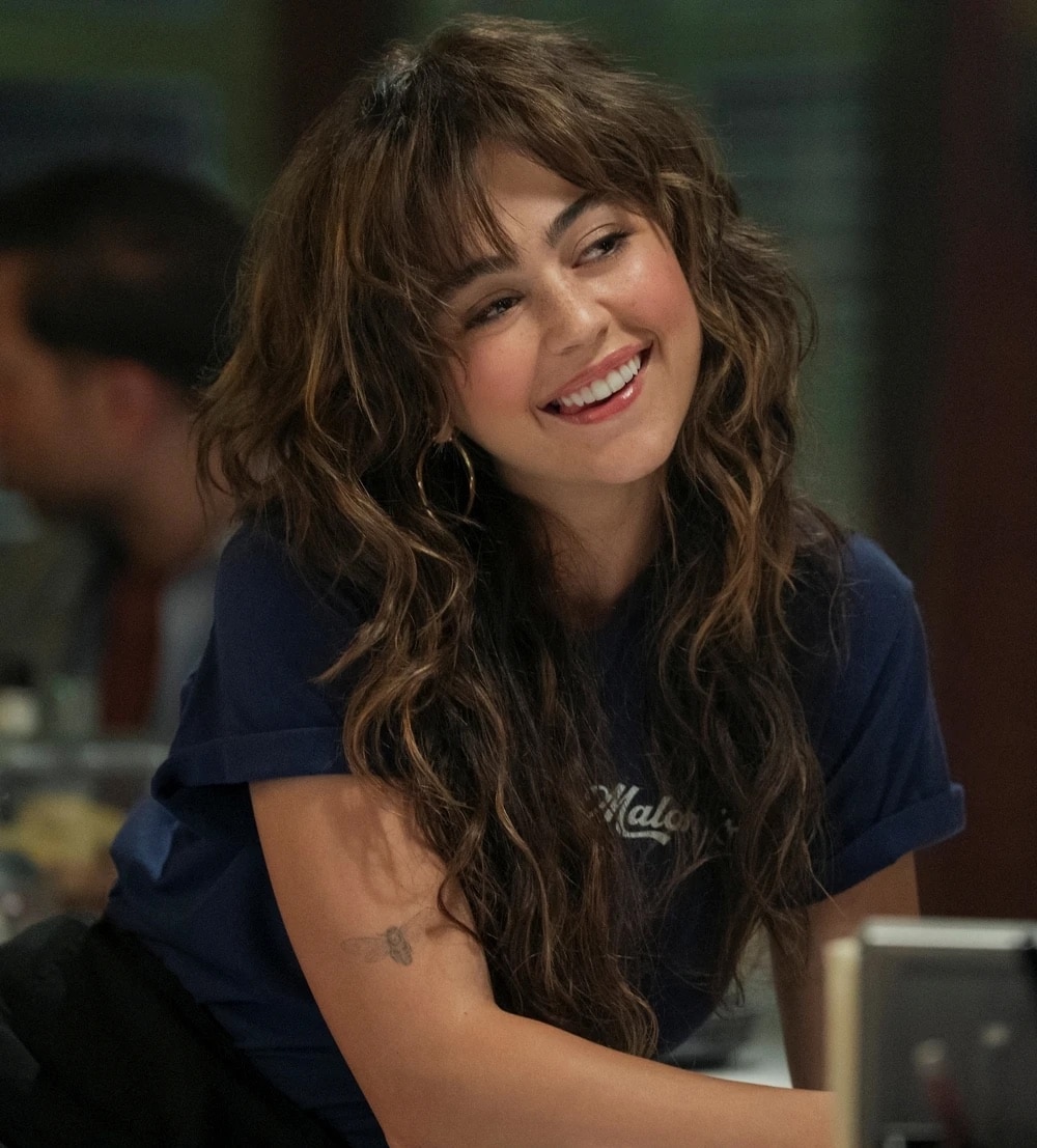 A young woman with long, wavy brown hair and bangs—reminiscent of top TV hairstyles—is smiling warmly. She is wearing a blue t-shirt and hoop earrings, with a small tattoo on her upper arm. The background is softly blurred.