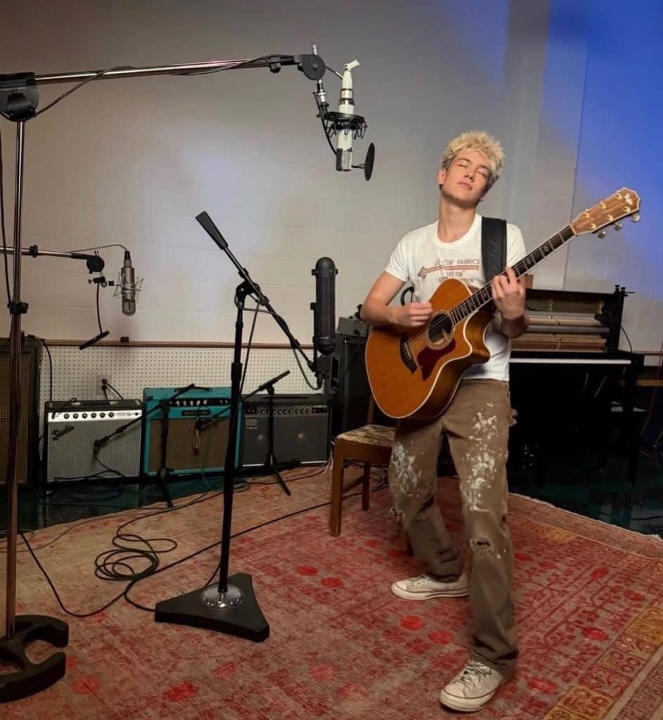 A person with short blond hair, resembling Hudson Stone, plays an acoustic guitar passionately in a recording studio, standing on a patterned rug surrounded by microphones and amplifiers. They wear a white t-shirt, paint-splattered pants, and white sneakers.