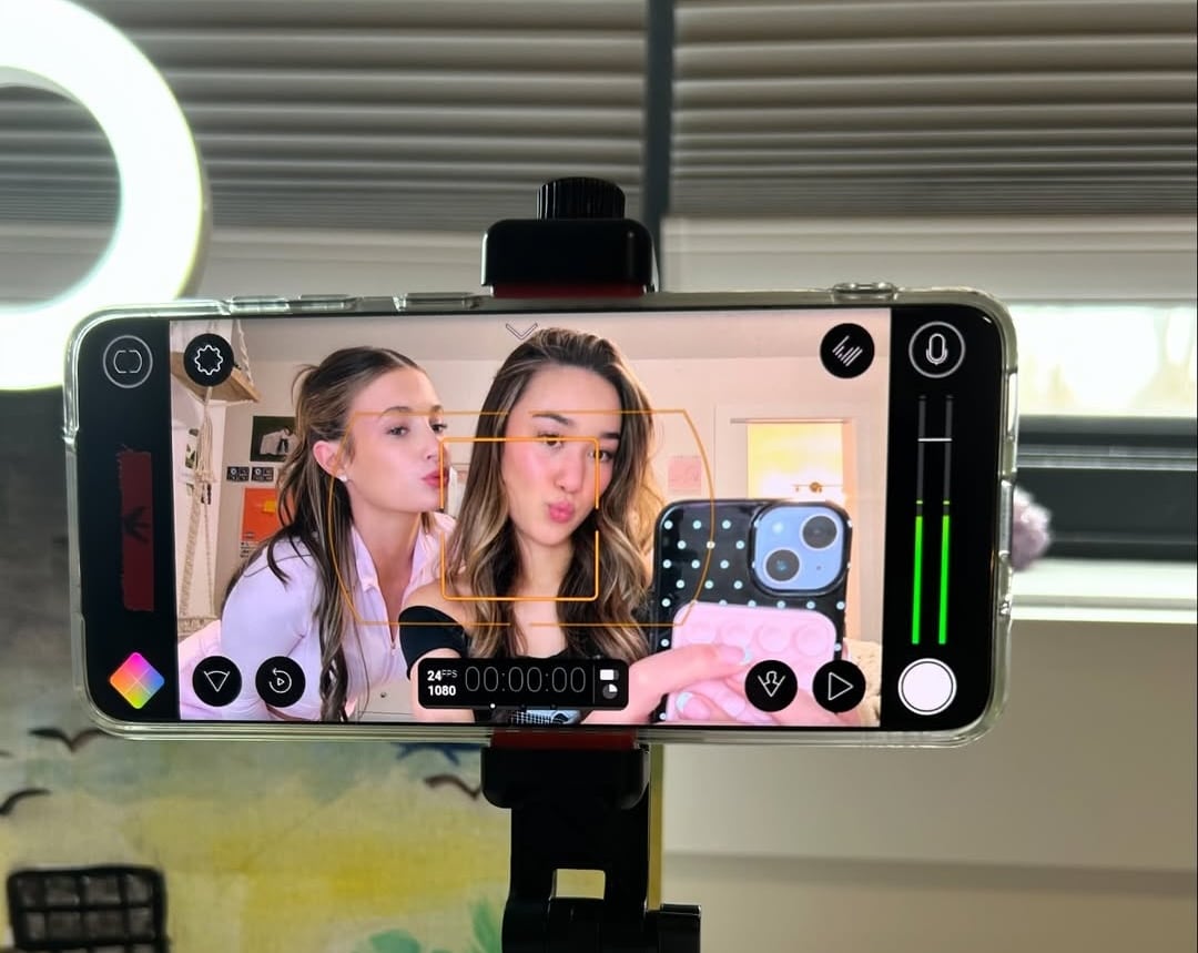 Two young women pose for a selfie in front of a smartphone on a tripod. The phone screen displays their image with camera app controls. A ring light, window, and an Ava Ro accessory are visible in the background.