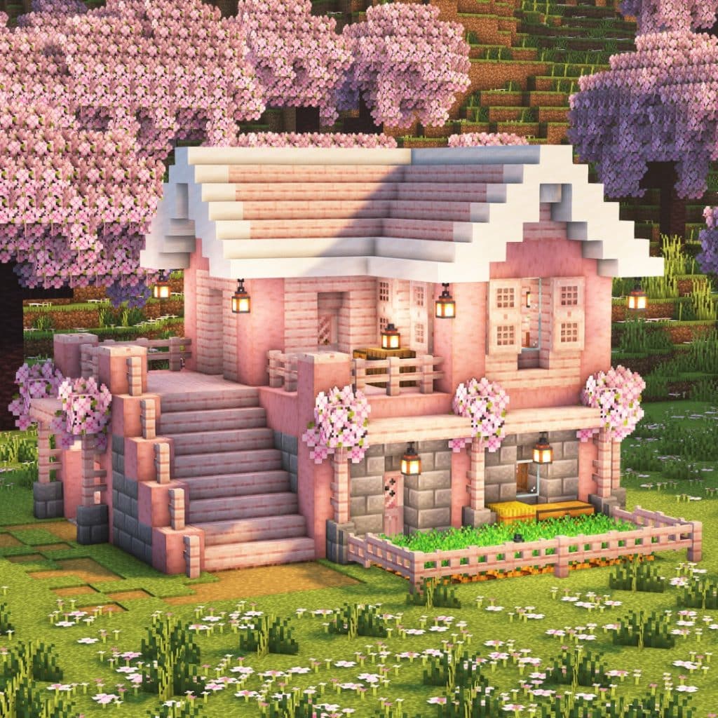 A two-story pink and white Minecraft house with lanterns, surrounded by cherry blossom trees and flowers, features a front porch, balcony, stone steps, and a small fenced garden.