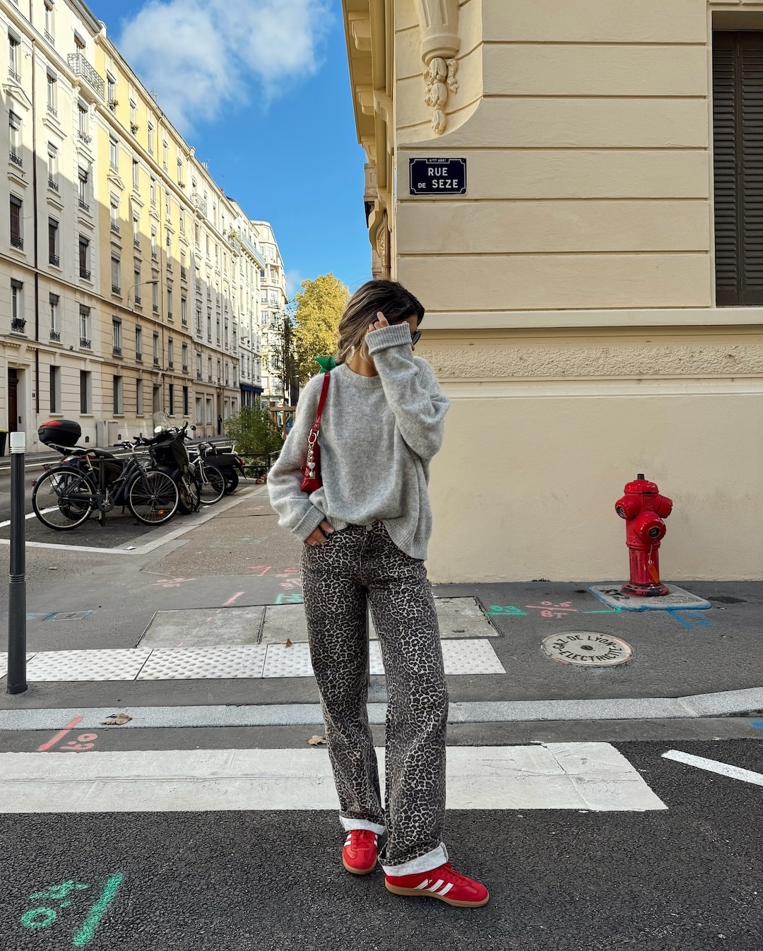 A person stands on a city street corner, covering their face. Sporting back-to-school shoes—red sneakers—they pair them with a gray sweater, leopard print pants, and a red bag. Bicycles, a fire hydrant, and buildings appear in the background under a blue sky.