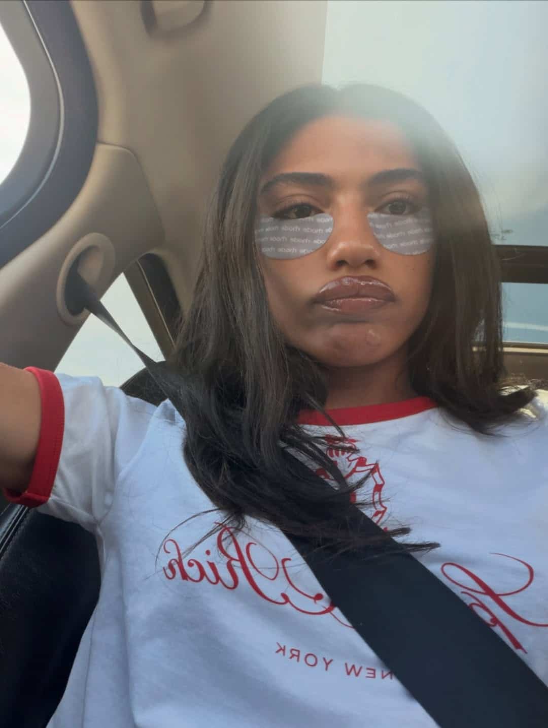 A woman with long dark hair wears under-eye patches and a white T-shirt with red trim, sitting in a car and looking at the camera with a pouty expression. Sunlight streams in from the window.