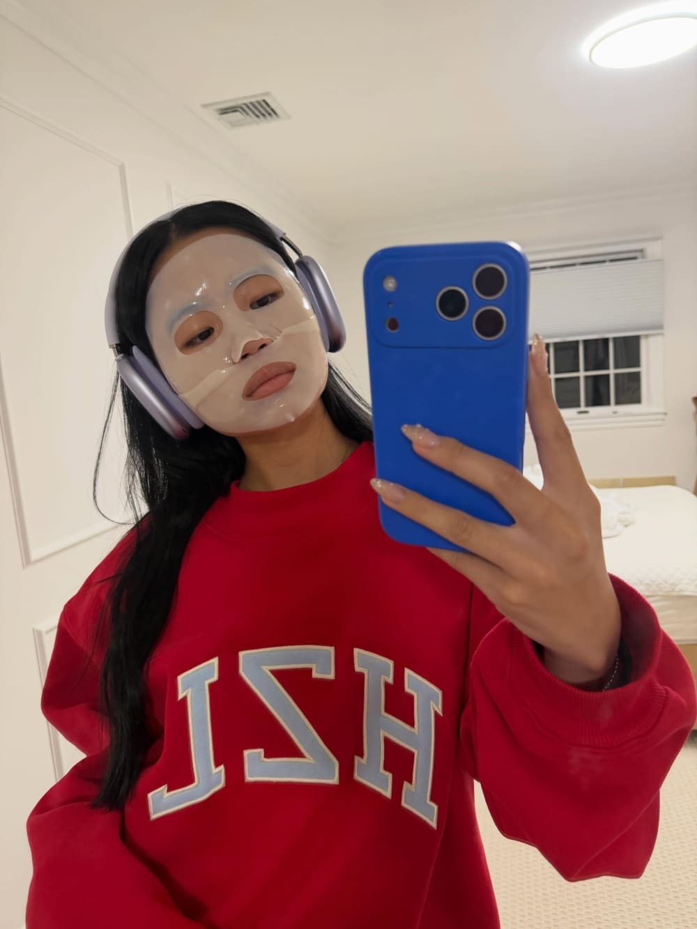 A person wearing a sheet face mask and headphones takes a selfie in a bedroom, wearing a red sweatshirt with large white letters.