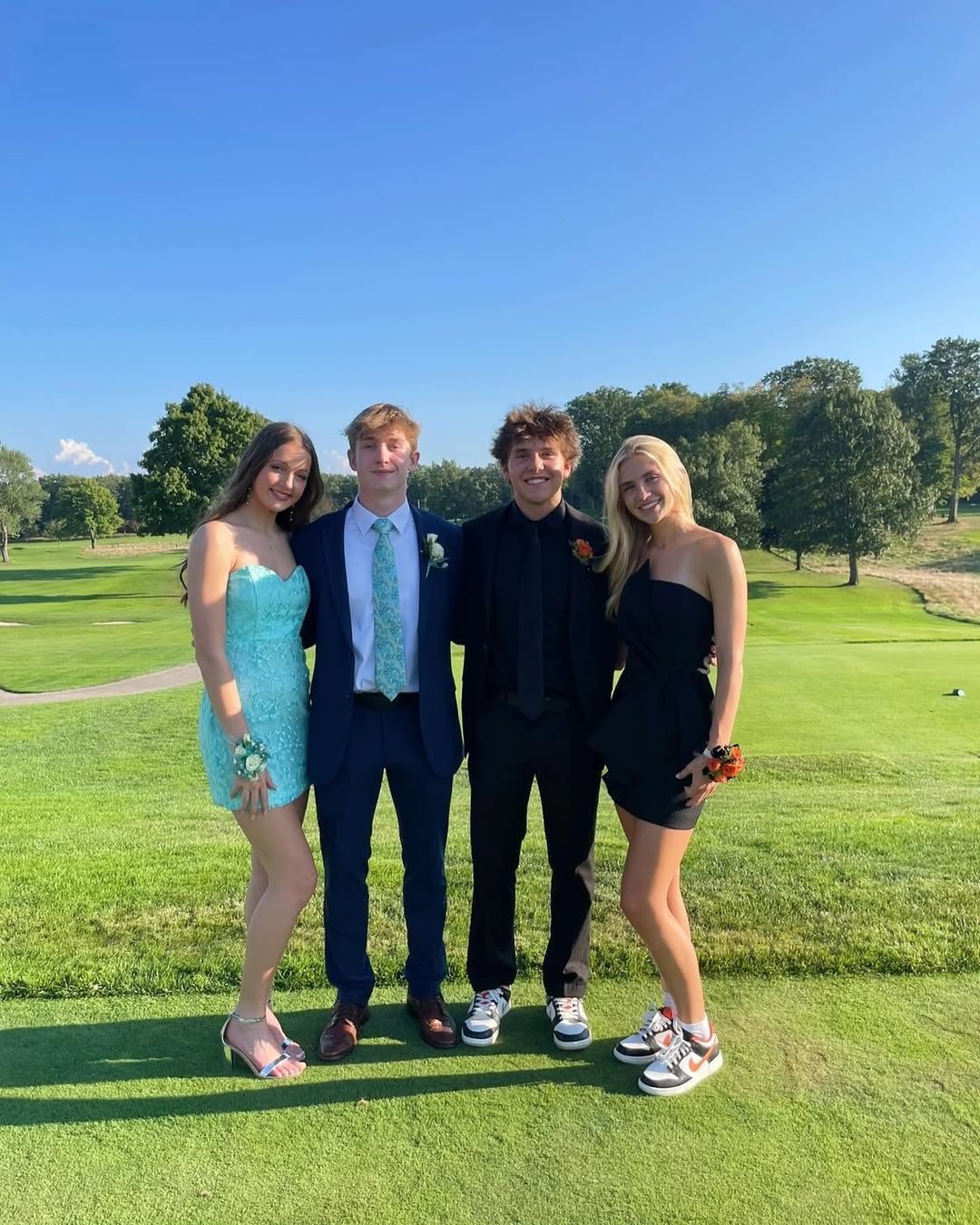Four young adults dressed in formal and semi-formal attire pose together on a sunny golf course. Two women wear dresses and corsages, while two men wear suits; all are smiling at the camera. Trees and green grass are in the background.