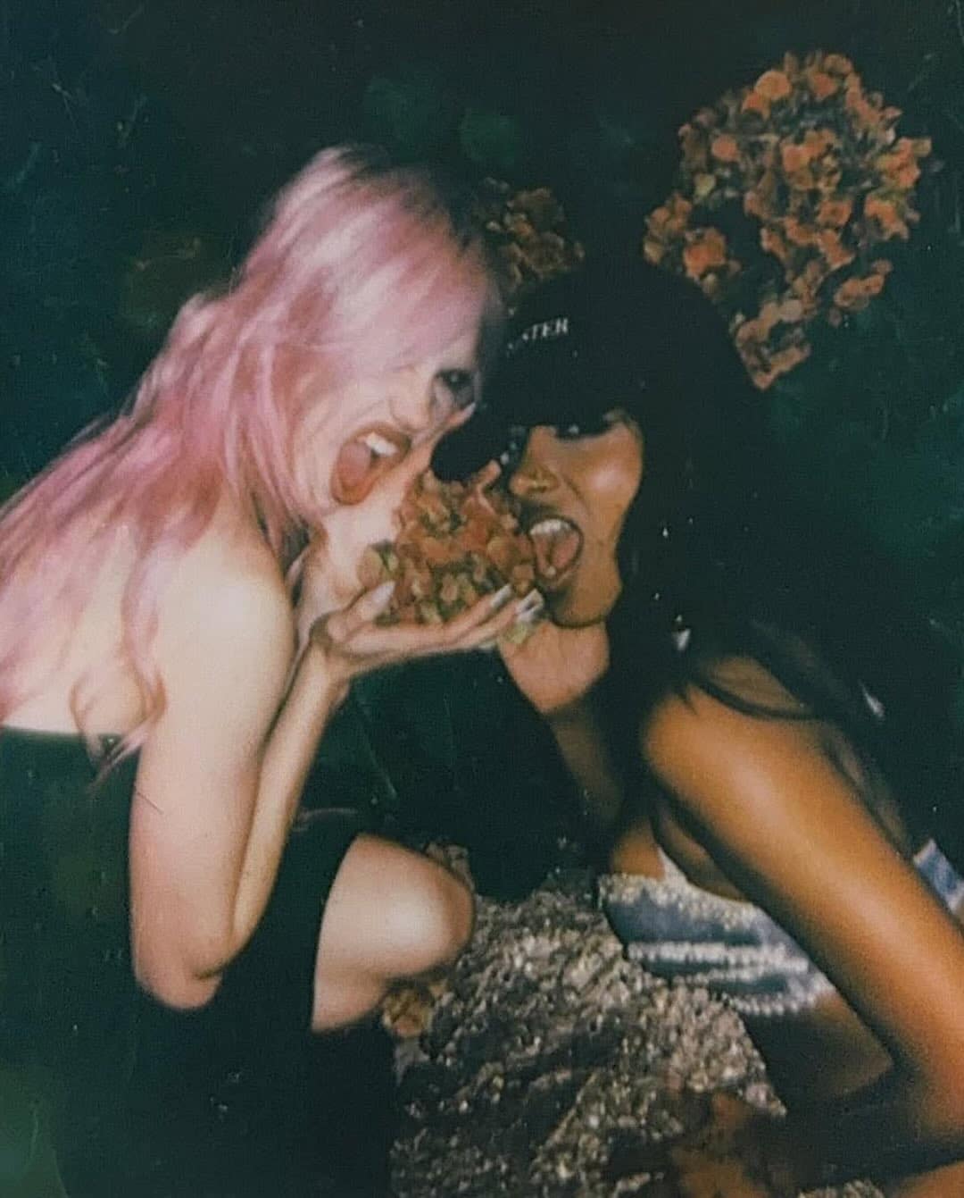 Two women pose playfully, appearing to bite a cluster of flowers. One has long pink hair, the other dark hair and a cap—radiating the energetic charm of a pop girl duo. Both are smiling, and the background is filled with more flowers. The photo has a candid, fun vibe that perfectly captures their pop duo personality.