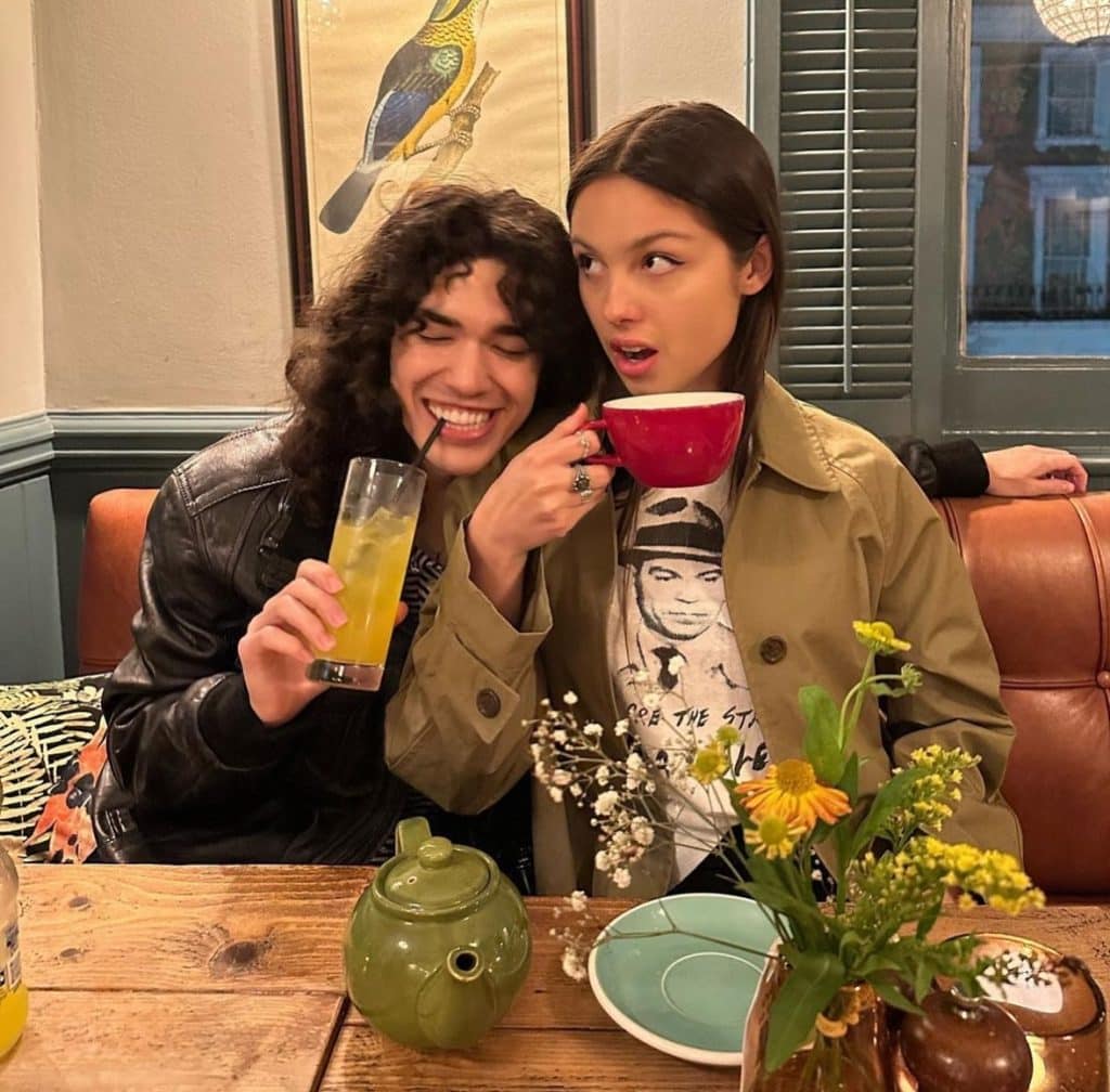 Two besties sit at a wooden table with a teapot, plates, and flowers. One person laughs holding a drink, while the other looks surprised holding a large red mug—just like a pop girl duo mid-quiz. A bird artwork hangs on the wall behind them.