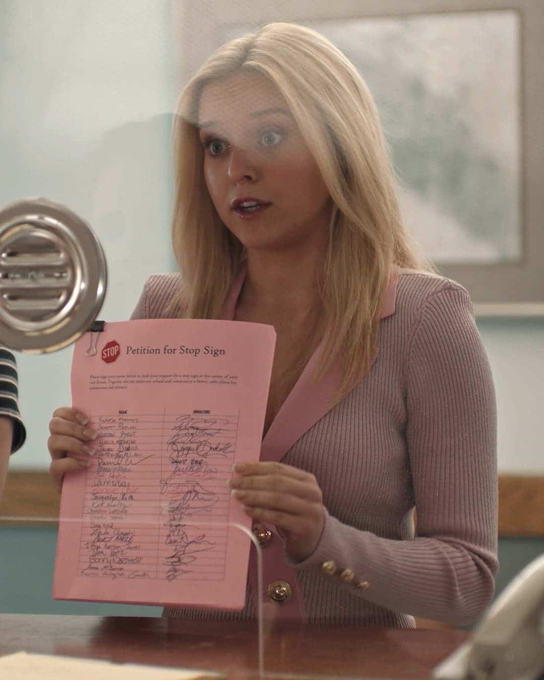 A woman with long blonde hair in a pink blazer holds up a pink petition form with signatures, standing behind glass at a counter, looking surprised or concerned—perfectly embodying Elle's guide to staying true to yourself and looking great, even in unexpected moments.