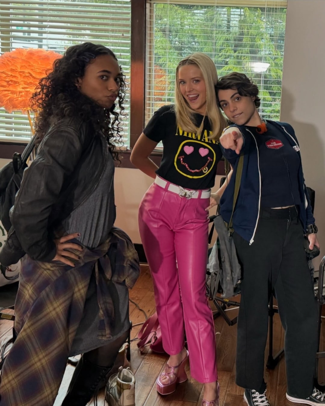 Three stylish people pose indoors, looking great and staying true to yourself. One wears a jacket and plaid shirt, another in a Nirvana tee and pink pants smiles, and the third in a blue jacket points at the camera. A bright orange lamp is in the background, perfectly capturing Elle's guide to effortless style.