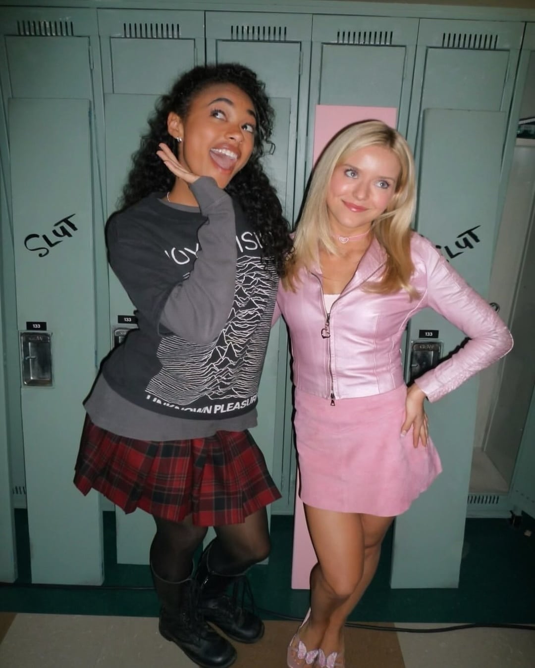 Two young women pose in front of green lockers, confidently looking great. One wears a black graphic sweatshirt and red plaid skirt; the other sports a pink top and skirt. The word “SLUT” is written on the lockers behind them, reminding us of the importance of staying true to yourself in every setting.