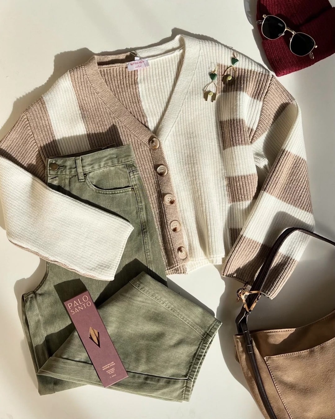 A beige and white striped cardigan, olive green jeans, brown purse, Palo Santo sticks, red beanie, and sunglasses are arranged on a white surface, lit by natural sunlight—everything you need to nix the back-to-school scaries in style.
