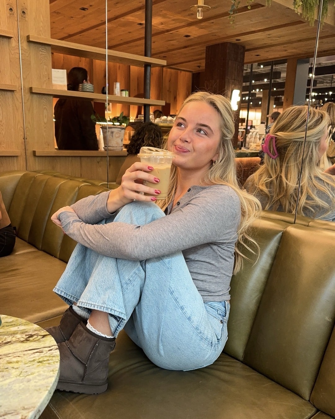 A woman with long blonde hair sits cross-legged on a green couch in a cafe, smiling and holding an iced coffee. She wears a gray top, blue jeans, and brown boots—looking relaxed and ready to conquer the Back-To-School Scaries with a positive outlook.