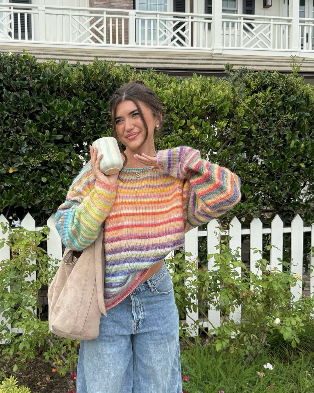 A woman wearing a colorful striped sweater and loose blue jeans stands in front of a white picket fence, holding a matching striped mug and smiling. With bushes and a house in the background, she looks ready to nix those Back-To-School Scaries with her cheerful style and positive energy.