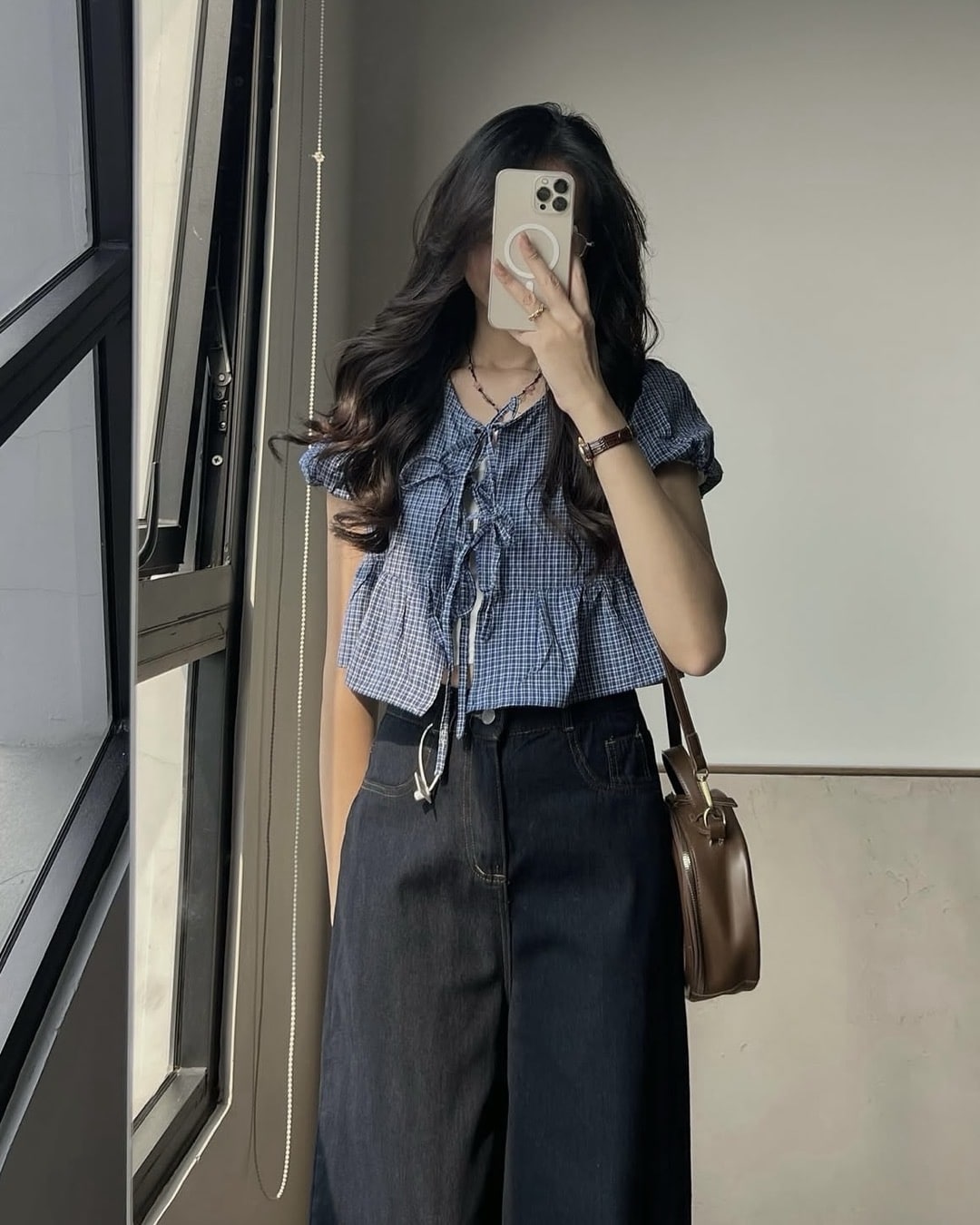 A person with long hair takes a mirror selfie, showcasing their unique style by wearing a blue checkered blouse and high-waisted dark pants, holding a phone in one hand and carrying a brown shoulder bag—an outfit that hints at thrift store shopping finds or even pieces inspired by BTS wardrobe looks.