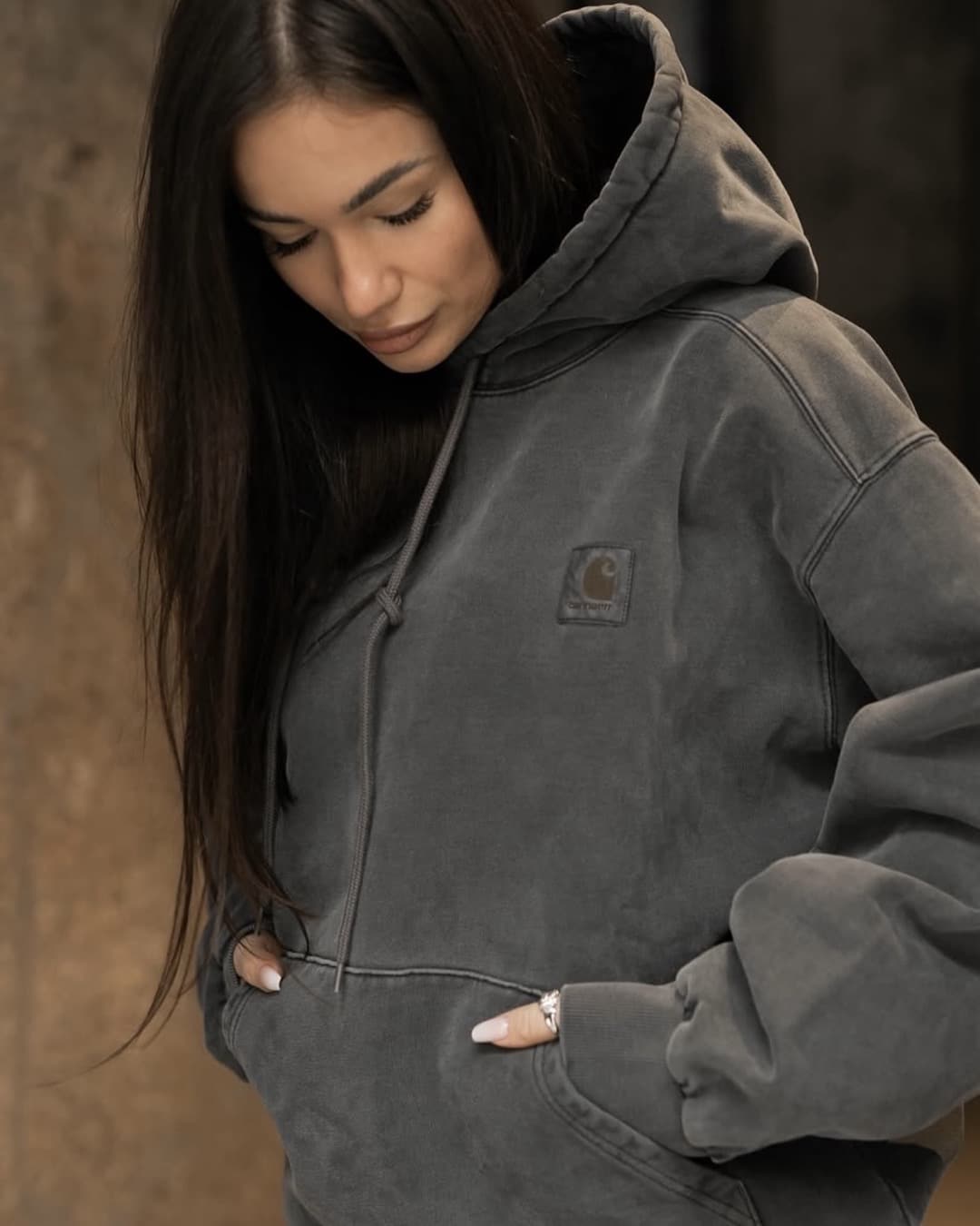 A woman with long dark hair looks down while wearing an oversized gray hoodie with a Carhartt logo, her hands in the front pocket—a casual style that suggests thrift store shopping. The background is blurred and neutral-toned.