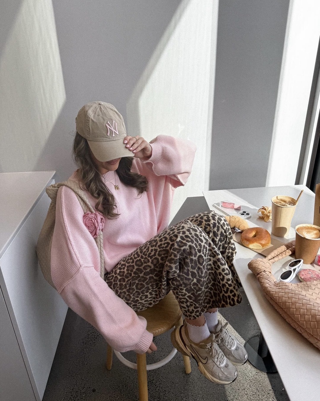 A person in a pink sweater, leopard print pants, white sneakers, and a beige NY cap—showcasing effortlessly cool Style inspired by BTS wardrobe trends—sits at a table with coffee, donuts, sunglasses, and a woven bag in a bright cafe.