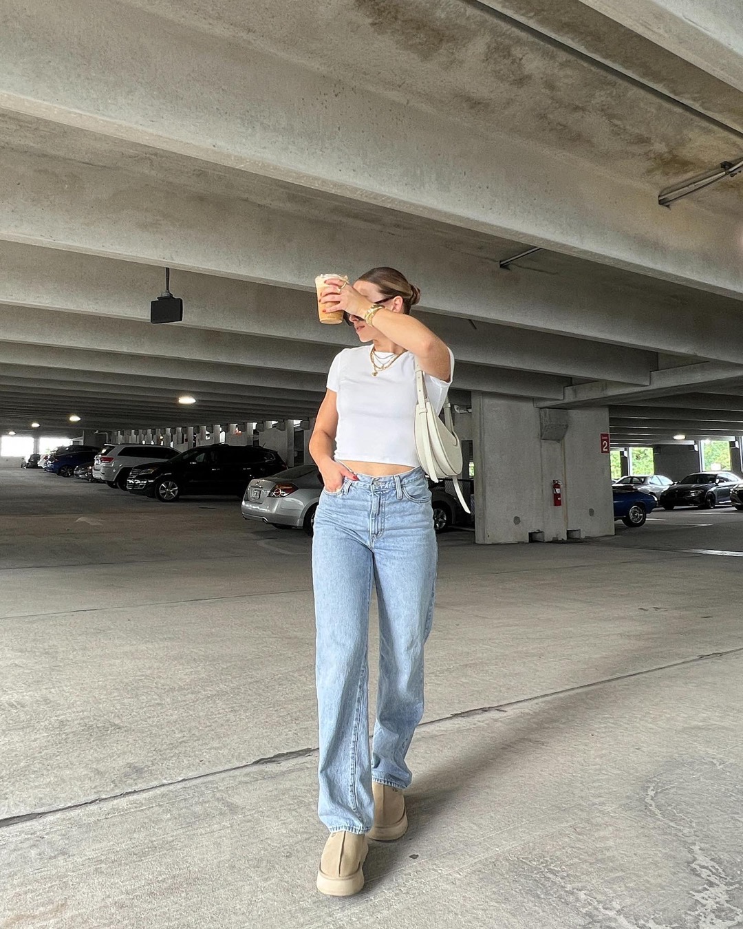Woman in a parking garage wears light blue jeans, a white fitted shirt, beige shoes, sunglasses, and holds an iced drink in front of her face—a look that could have easily come from a curated thrift store shopping list. Cars are parked in the background.