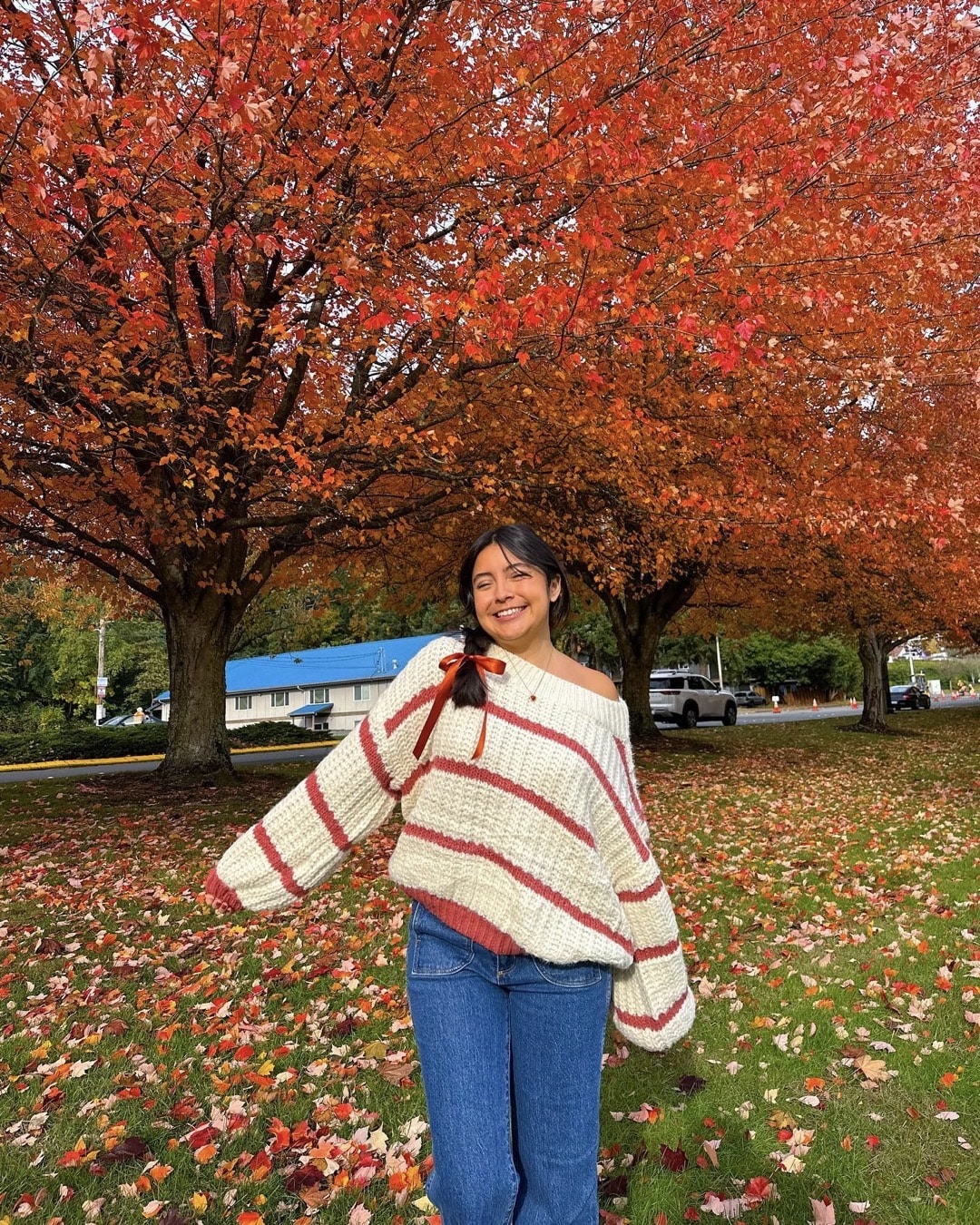 A woman in a striped sweater and jeans stands smiling under vibrant orange autumn trees, surrounded by fallen leaves on the grass, showcasing her effortless style that hints at BTS wardrobe inspiration and a love for thrift store shopping.