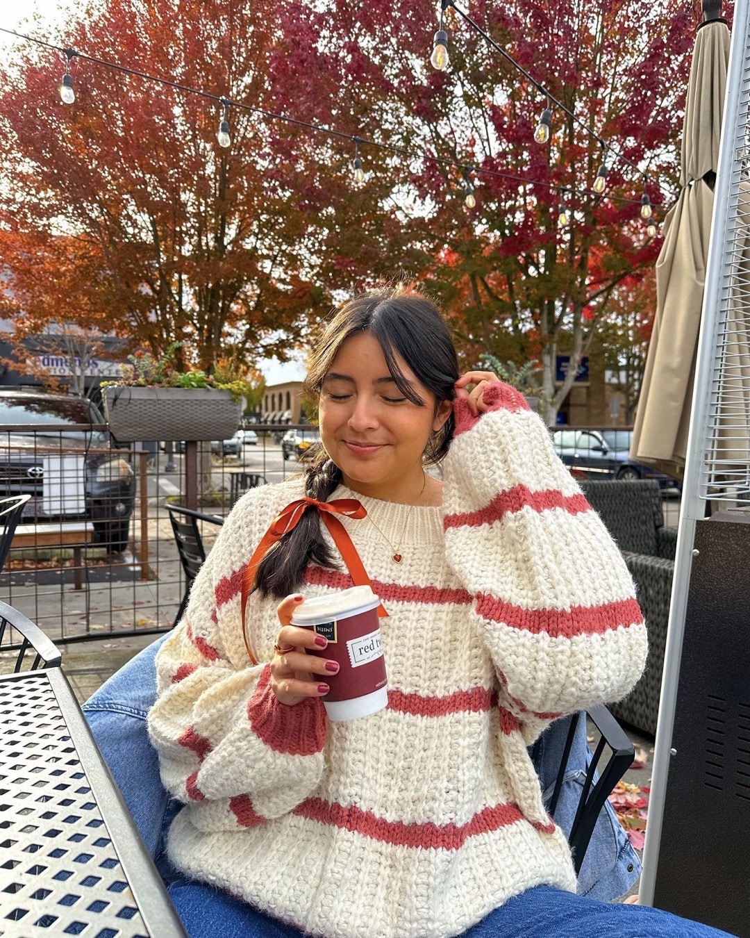 A woman in a cozy striped sweater—perhaps a cherished find from a thrift store—sits outdoors, smiling with closed eyes and holding a coffee cup. Autumn trees with red leaves and string lights decorate the background.