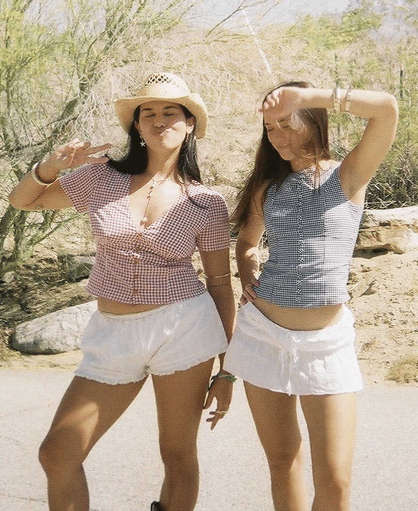 Two women, celebrating an end-of-summer reunion, pose outdoors in stylish summer outfits with white shorts and checkered tops. One wears a hat and flashes a peace sign, while the other raises her arm—both smiling joyfully in the bright sunlight. Desert plants set the scene for this unforgettable BFF reunion.