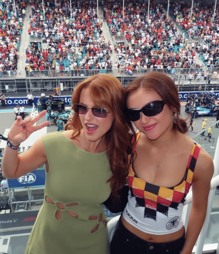 Two women in sunglasses pose and smile at a crowded racetrack, one making a peace sign. Capturing the spirit of an unforgettable BFF reunion, the background shows stands filled with spectators and part of the track with event staff visible.