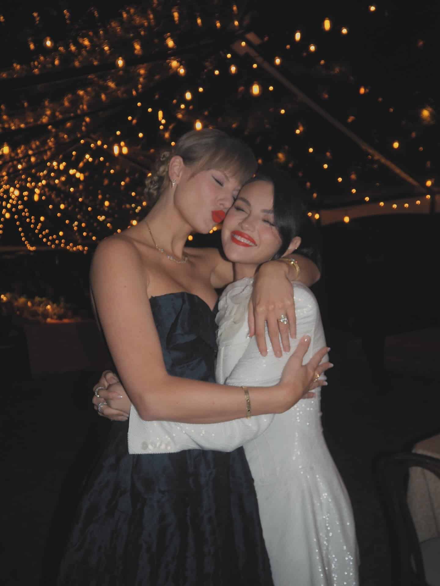 Two women dressed formally hug and smile under a canopy of warm string lights at night. Channeling the energy of a pop girl duo, one woman wears a black dress and kisses the other's cheek; the other wears a white dress and smiles.