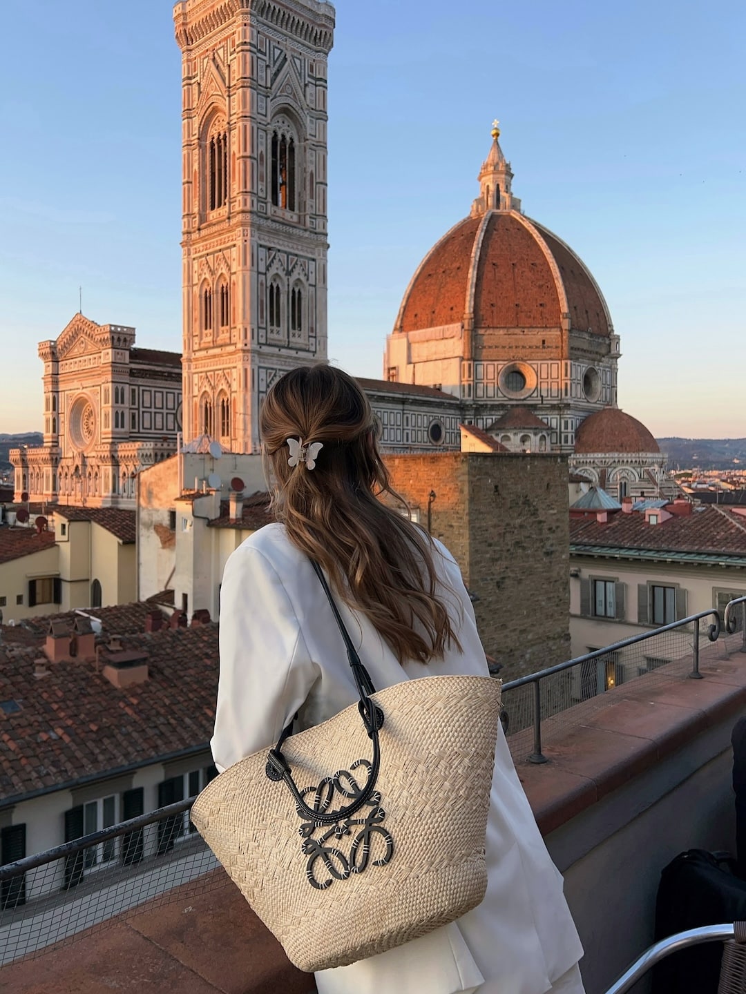 A woman with wavy brown hair, wearing a white outfit and carrying a straw bag, stands on a rooftop terrace overlooking Florence’s Duomo and the Campanile at sunset.