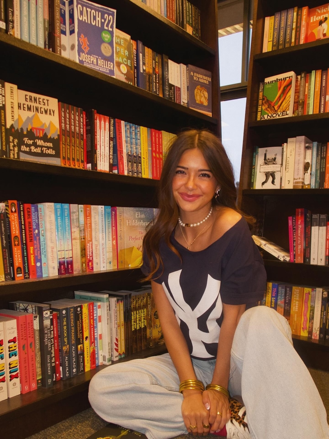 A young woman with long brown hair, wearing a dark blue Yankees shirt and light pants, sits smiling on the floor in front of colorful bookshelves in a bookstore.