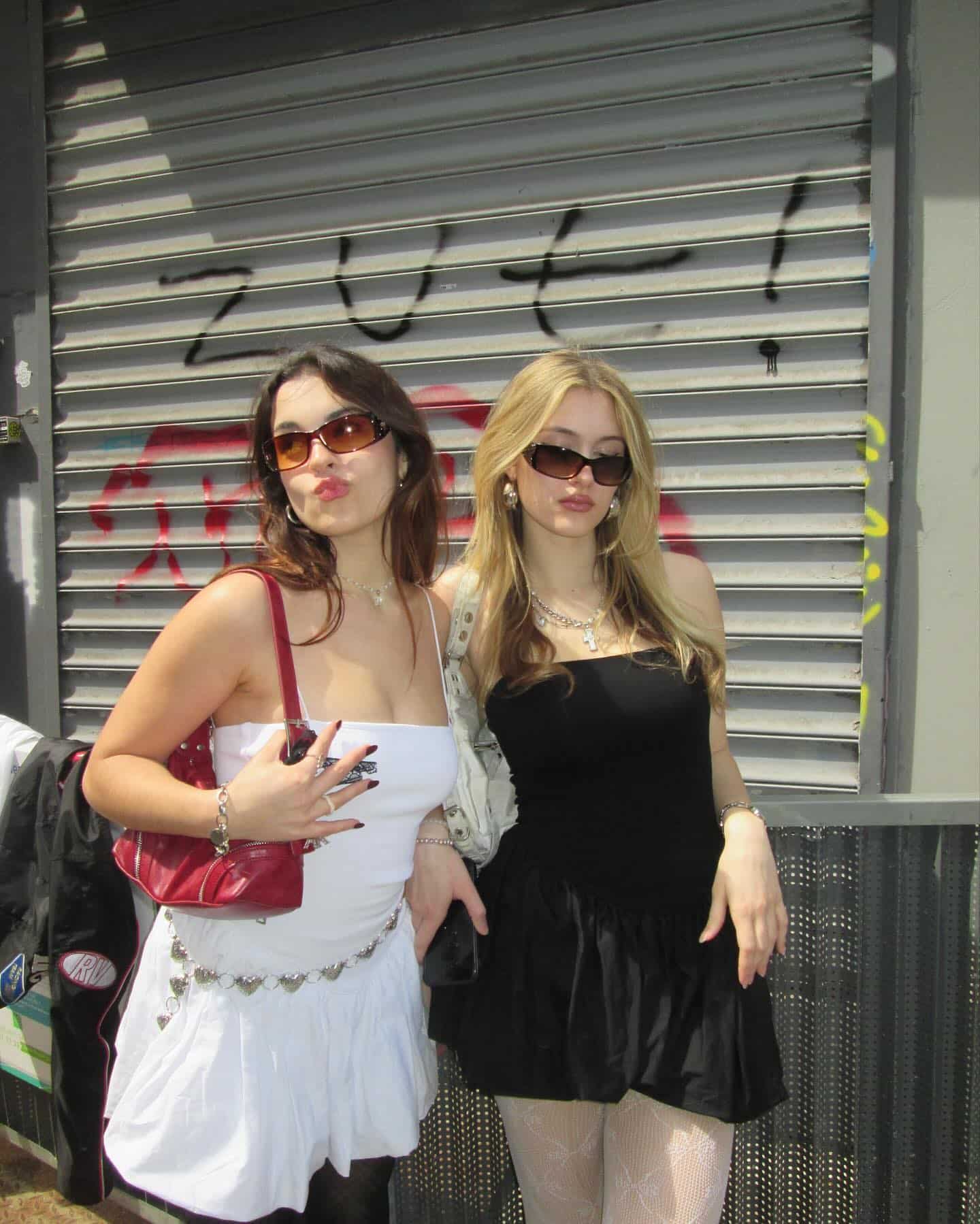 Two women pose in front of a metal shutter with graffiti, capturing the essence of an End-Of-Summer Reunion. Both wear sunglasses and mini dresses—one in white, the other in black. They hold small purses and make a pouty face for the camera, perfectly embodying BFF vibes and celebrating their friendship.