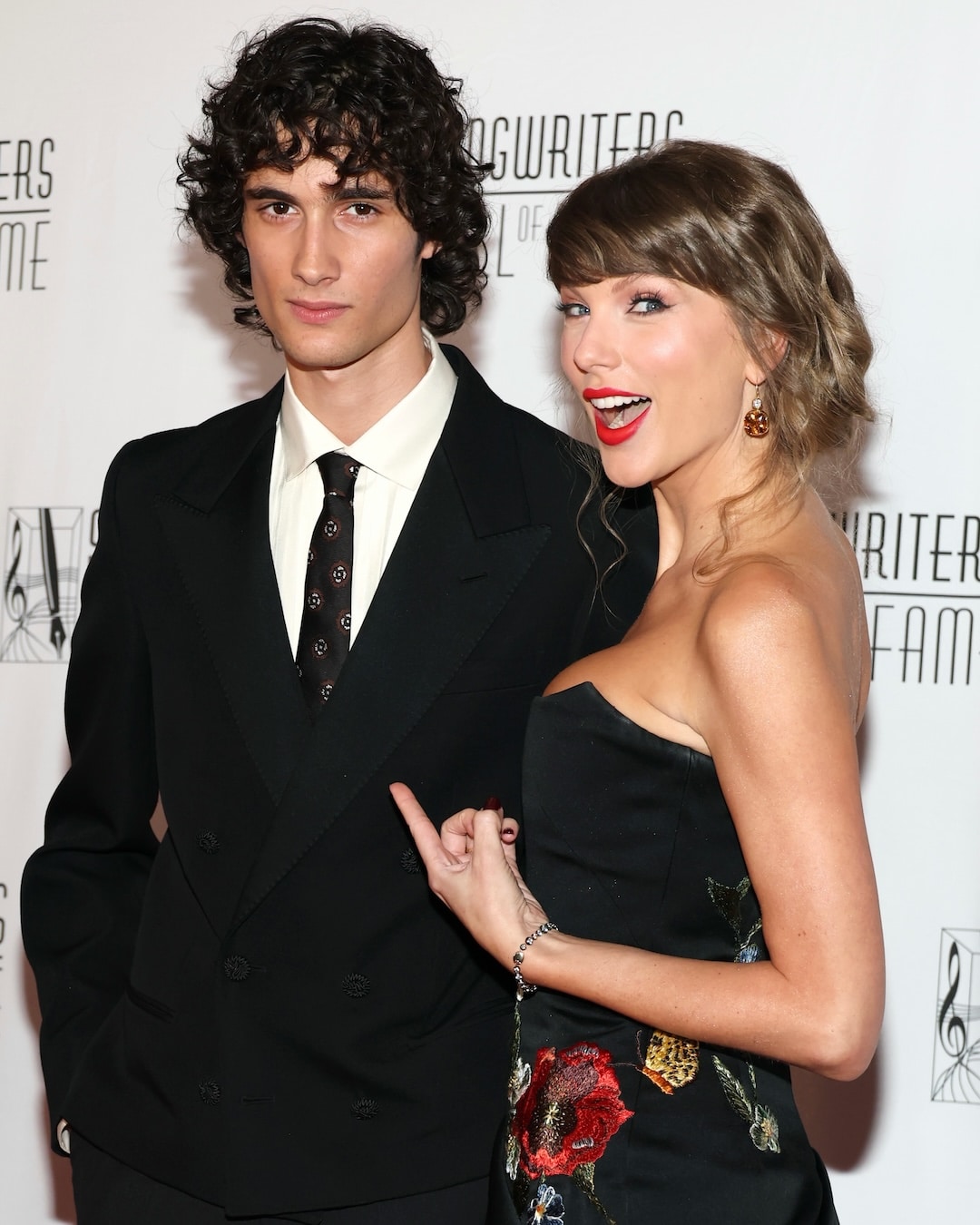 Two people dressed formally pose together on a red carpet, standing in front of a backdrop that reads Songwriters Hall of Fame. One person smiles and points at the other while the other maintains a neutral expression.