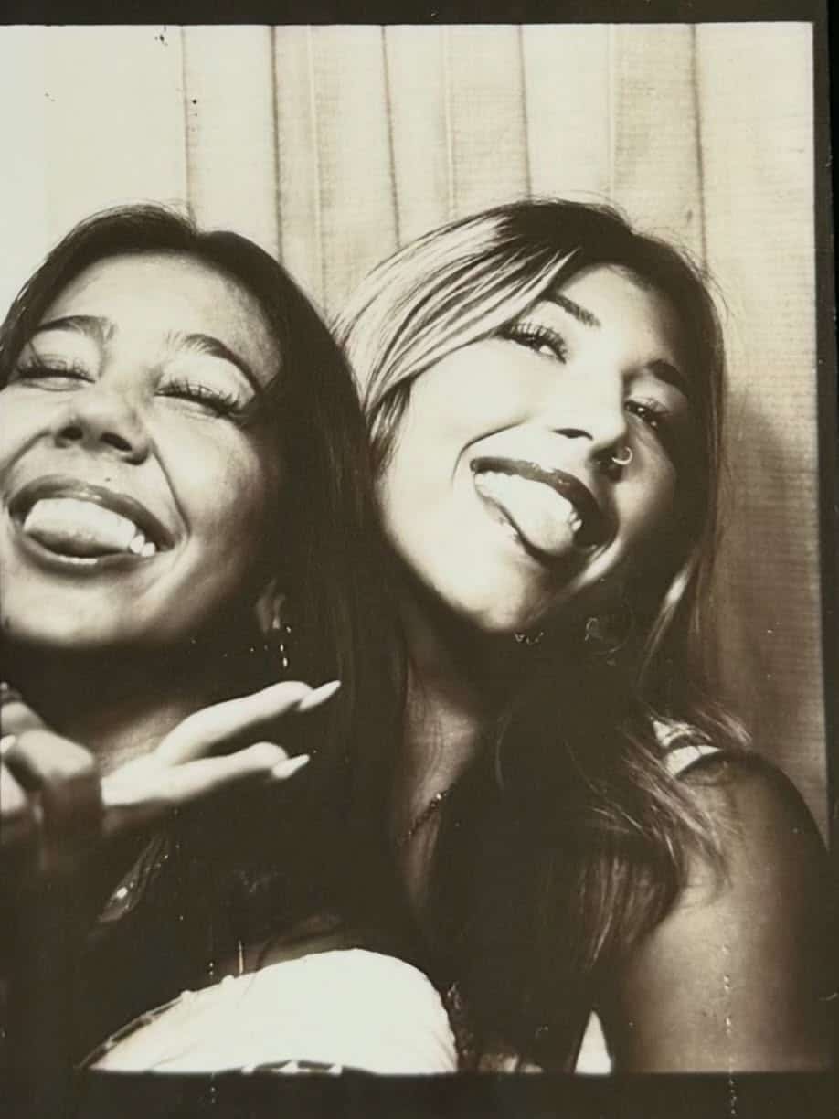 Two young women, caught in a playful pose for a black and white photo booth picture, smile and stick out their tongues—capturing the joy and carefree spirit of an end-of-summer reunion. Both appear delighted, celebrating summer friendship in this candid moment.