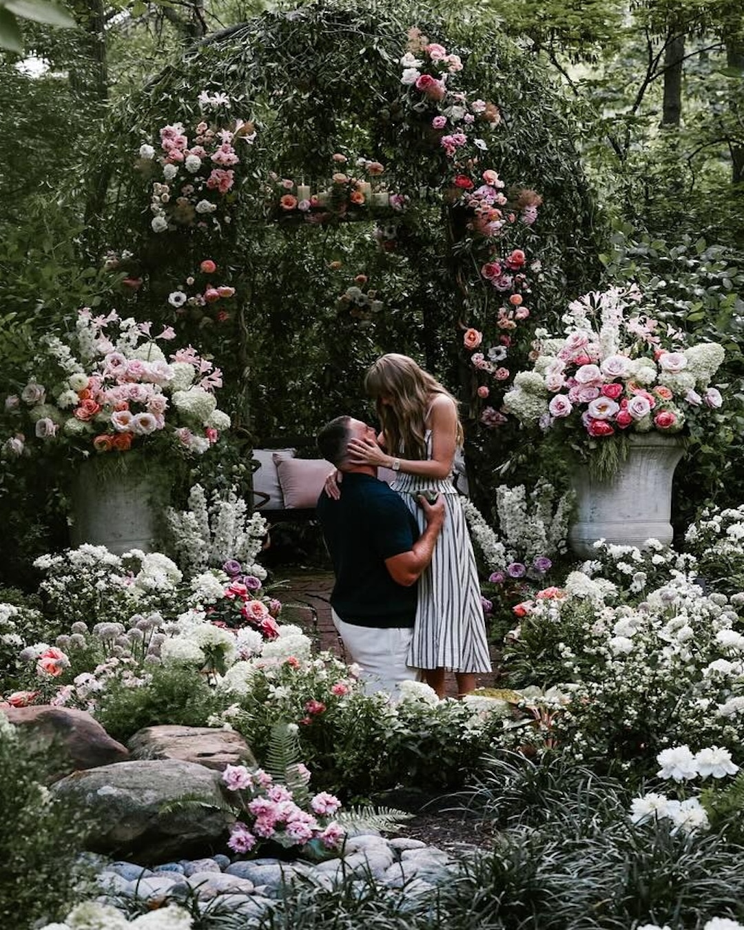 A man kneels before a woman, holding her waist, as she touches his face in a lush garden filled with white and pink flowers. An arched floral arrangement and two large vases frame the romantic scene, evoking Taylor Swift’s Married Era magic.