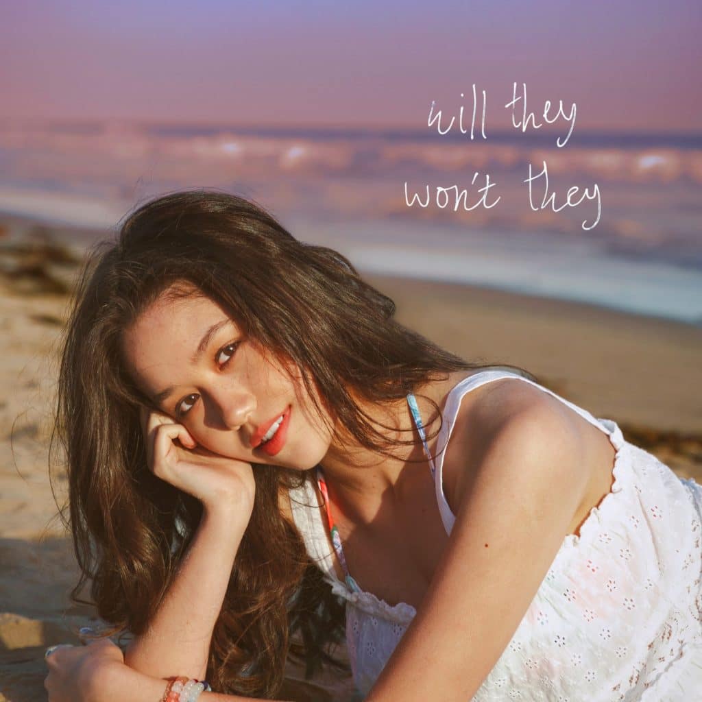 Young woman with long hair in a white dress lies on a sandy beach, smiling at the camera. The ocean is in the background, and the words will they wont they are written in white text. There’s an air of bliss around her, capturing a moment of pure joy.