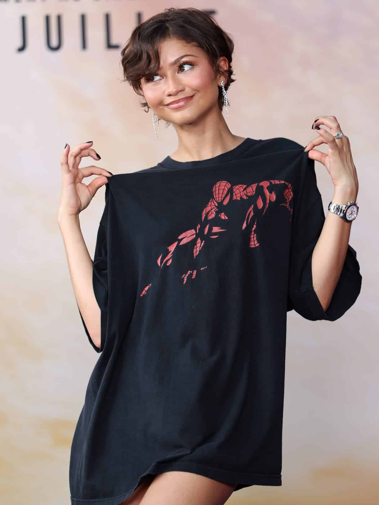 A woman with short curly hair smiles while holding out the sides of an oversized black T-shirt with a red SpiderMan graphic. She is wearing dangling earrings and a watch, channeling iconic looks in front of a light-colored background.