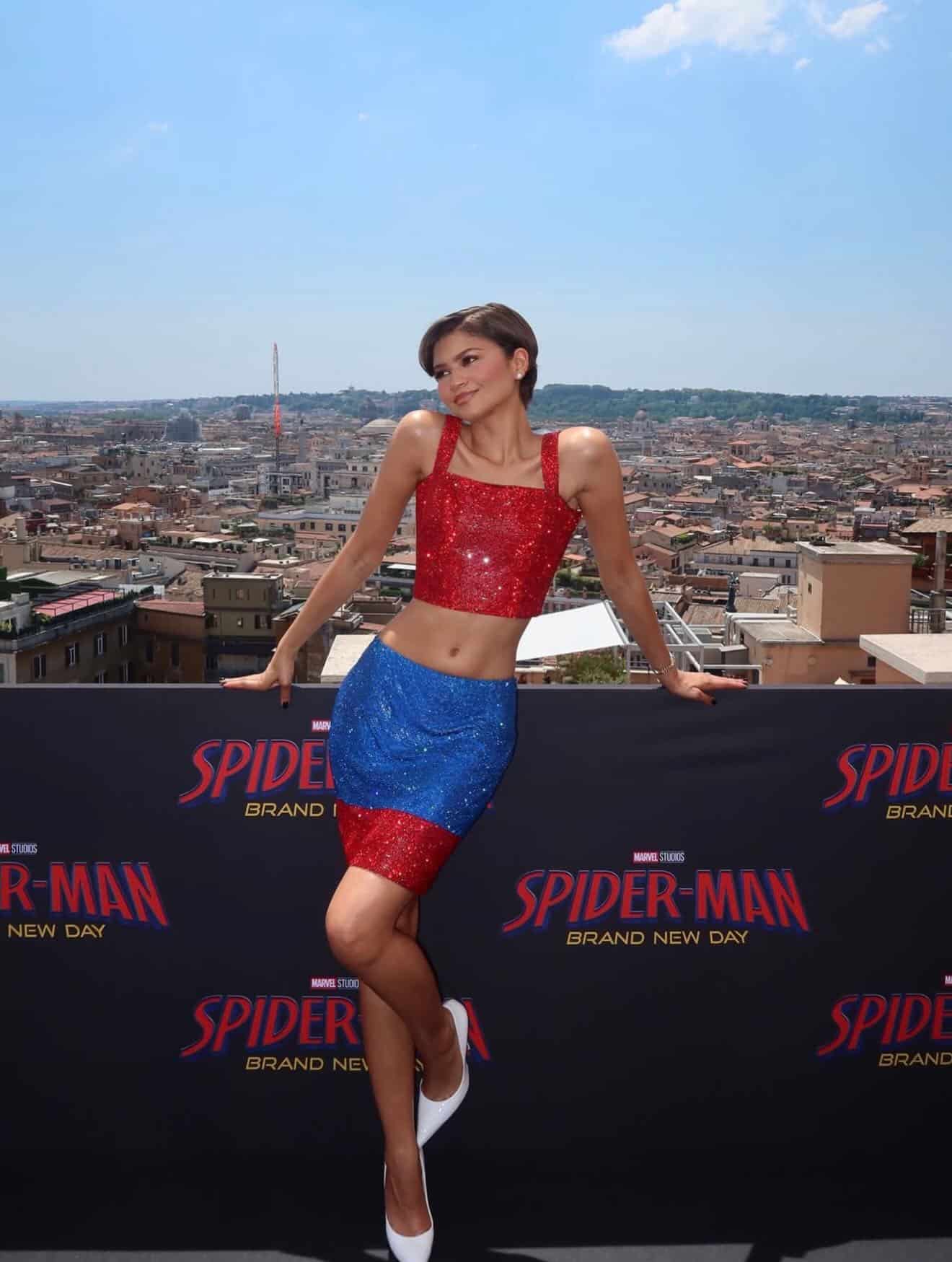 Zendaya showcases one of her iconic looks in a glittery red and blue two-piece, posing on a rooftop with a Spider-Man: Brand New Day banner set against the cityscape under the sunny sky.