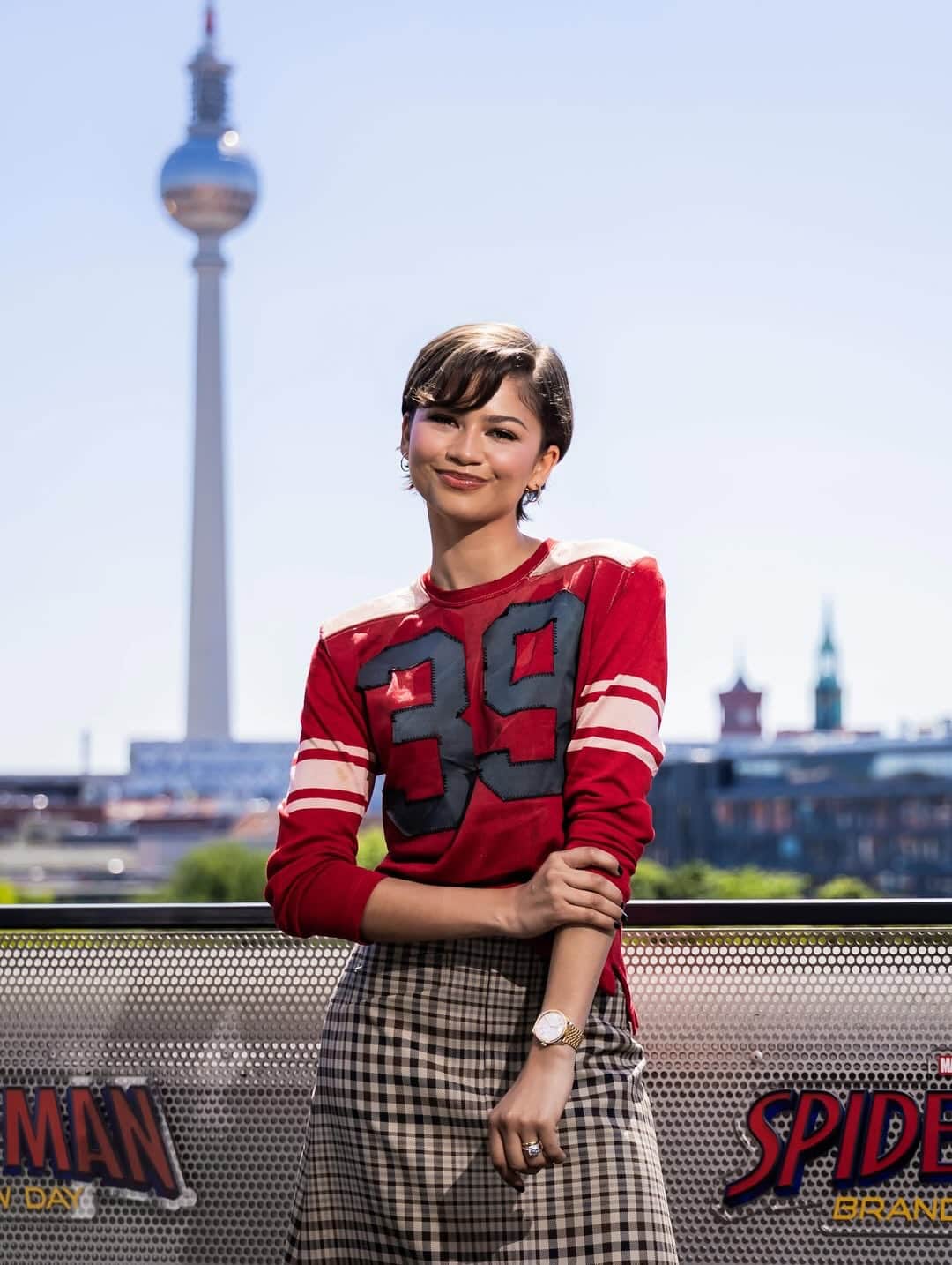 A young woman in a red sports jersey and plaid skirt stands outdoors, smiling in front of a cityscape with a tall tower. The railing features SpiderMan: Far From Home logos, channeling one of Zendaya’s iconic looks from the film.