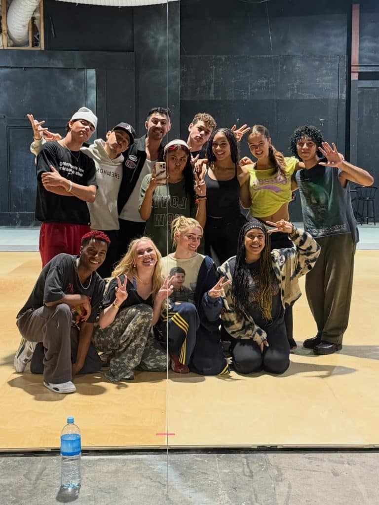 A group of 13 people pose and smile in a dance studio, some crouching and others standing, making peace signs. There’s a water bottle on the floor in front. A mirror behind them splits the image down the middle, giving the scene a lively vibe reminiscent of cast photos from shows like Zombies 5: Secrets of the Sea.