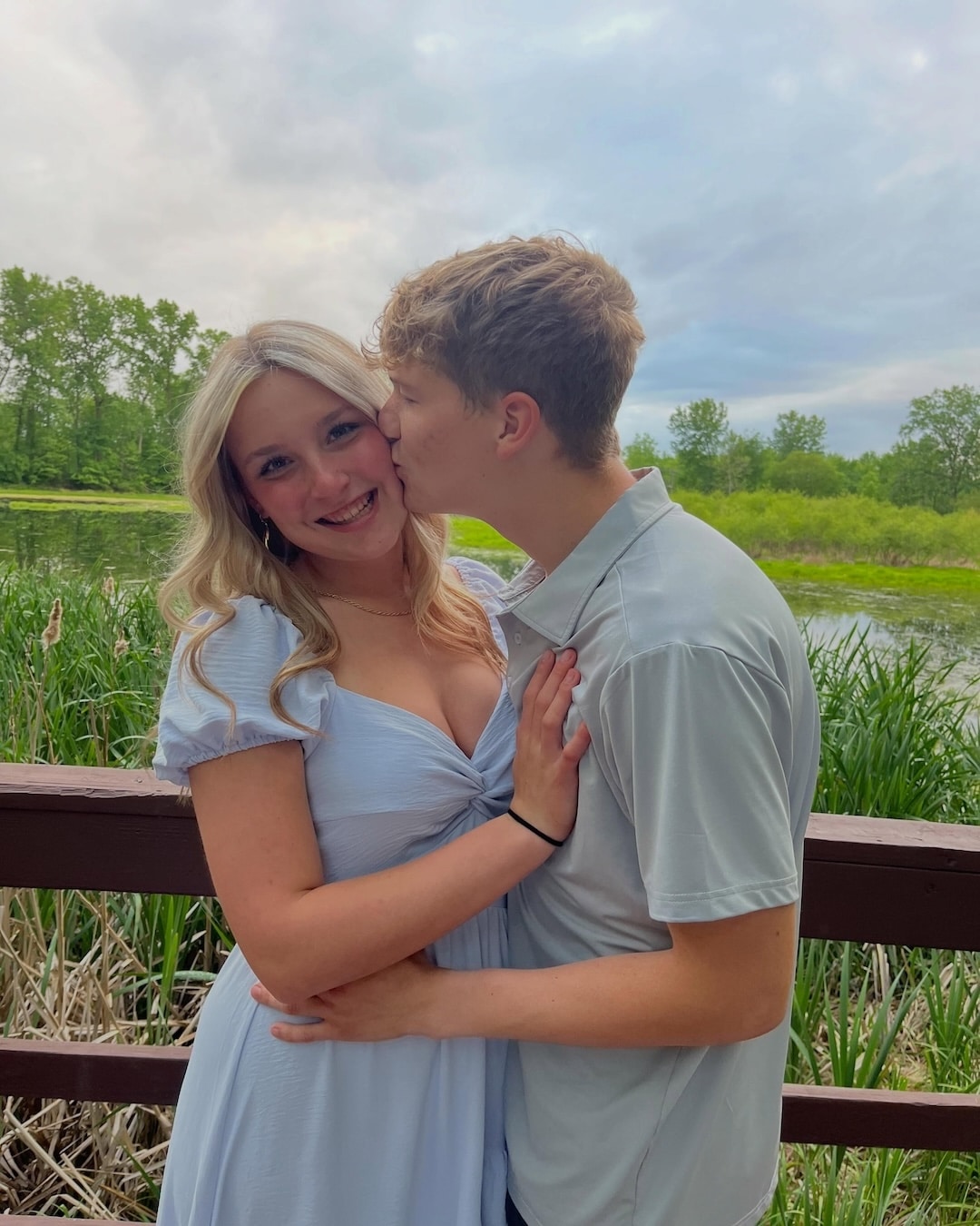A young couple stands on a wooden bridge, smiling; the man kisses the woman on the cheek. They wear light clothing, and a lush green landscape hints at a summer crush as trees and water create a romantic backdrop.