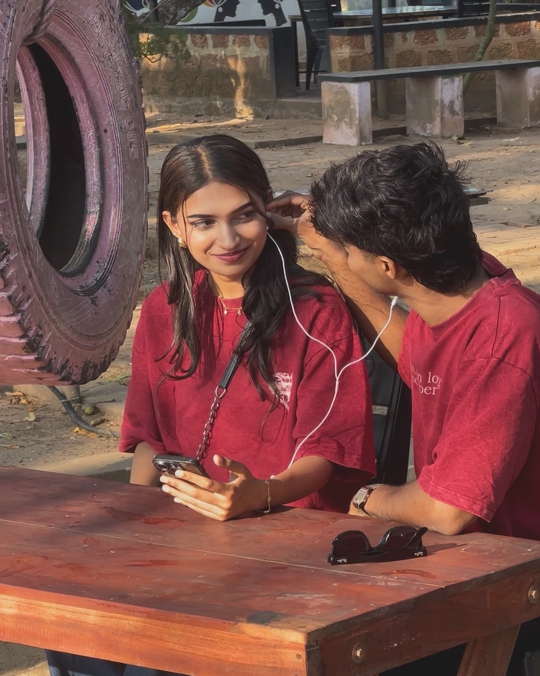 A young woman and man, both in matching red shirts, sit at a wooden table outdoors sharing earphones and smiling—summer crush vibes. The woman holds a phone while sunglasses rest on the table and a nearby tire swing adds to the carefree mood.