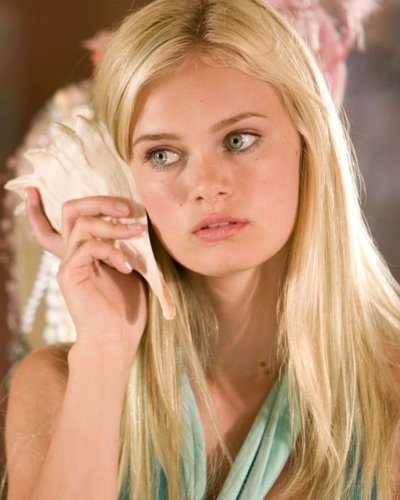 A young woman with long blond hair holds a large seashell to her ear, looking thoughtfully to the side. She wears a light blue top and is set against a softly blurred background.