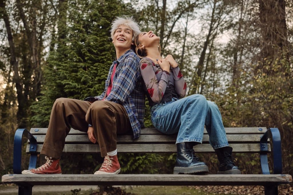 Two people sit back-to-back on a park bench, laughing and looking up. Dressed in casual, colorful clothes and boots, they capture the playful spirit of camp rock 3, with trees and greenery in the background enhancing this joyful outdoor moment.