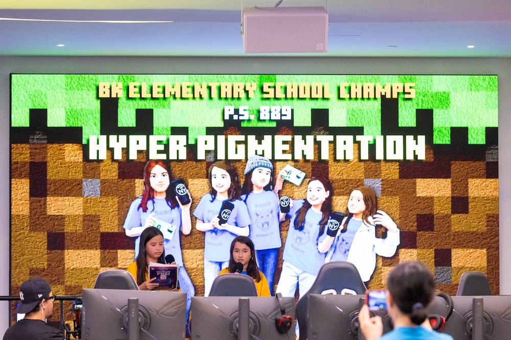 Six students in blue shirts pose onstage in front of a Minecraft-themed screen reading BK Elementary School Champs P.S. 889 Hyper Pigmentation, proudly showcasing their skills as they climb the leaderboard, while others sit at computers in the foreground.