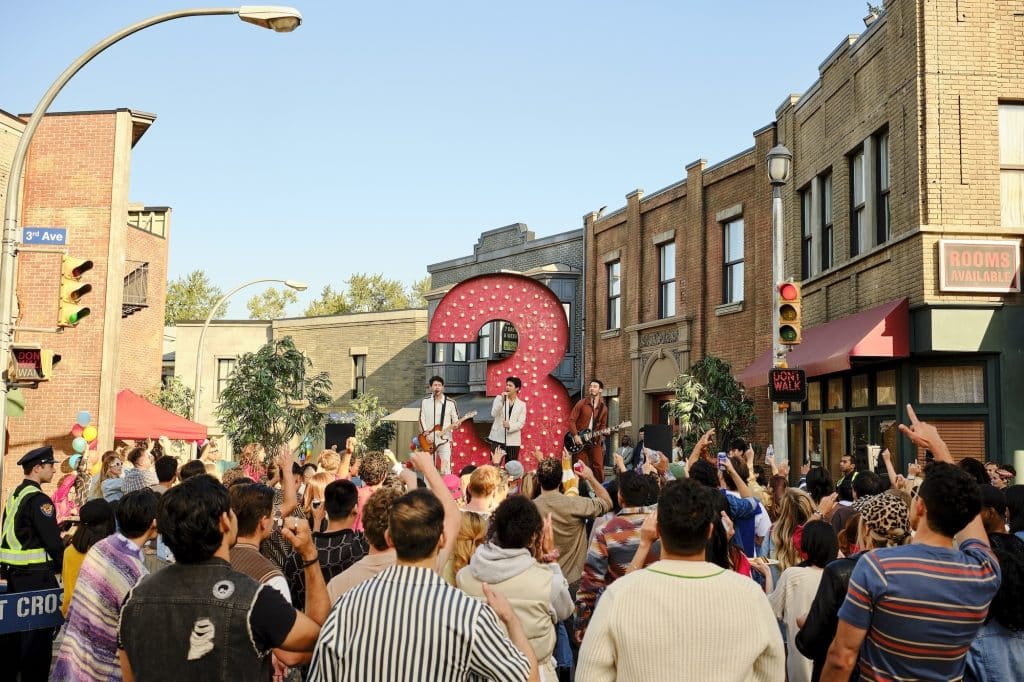 A band performs on an outdoor street stage in front of a large illuminated number 3, surrounded by a cheering crowd, creating an atmosphere reminiscent of Camp Rock 3. Buildings and a police officer are visible in the background.