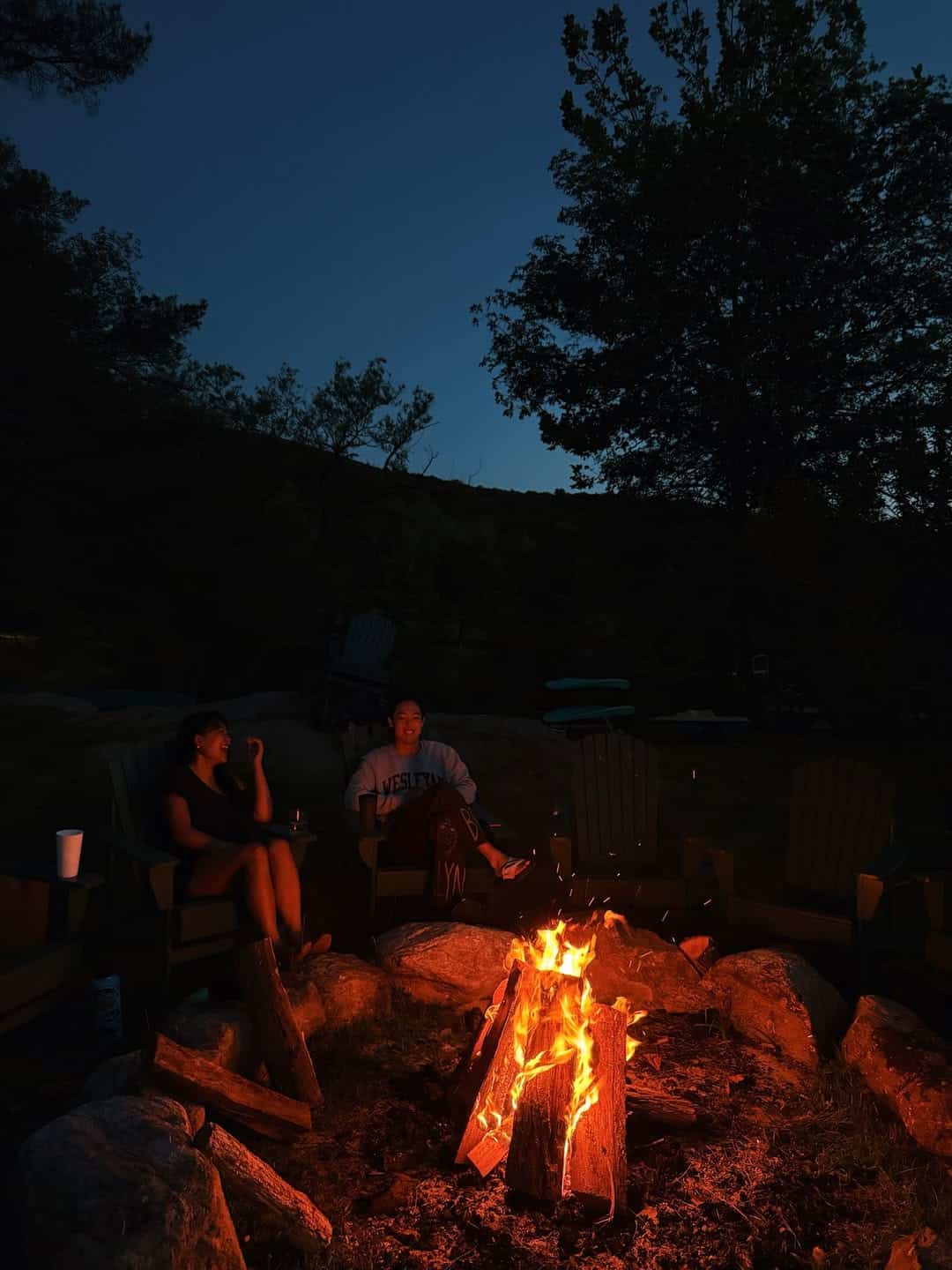Two people sit in chairs around a campfire at dusk, with trees and hills in the background. The fire glows warmly, illuminating their faces against the dark evening sky.