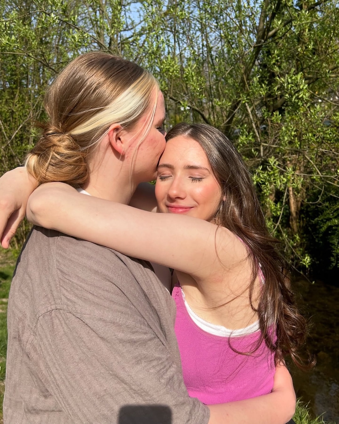 Two young women are hugging outdoors on a sunny day, sharing smiles and warmth. Green trees and sunlight fill the background, capturing the joyful spirit of a summer crush in this happy moment.