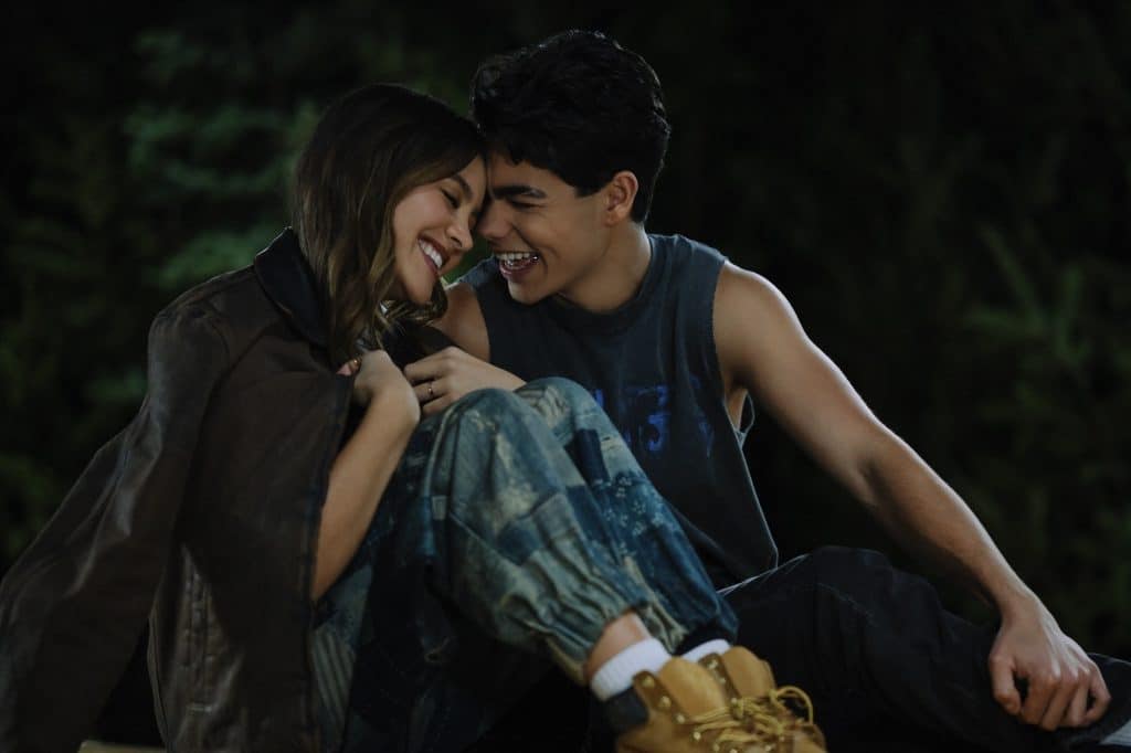Two young people sit close together at night, smiling and laughing as if they’re sharing a secret moment between takes on the set of Camp Rock 3. One wears a brown jacket and patterned pants, the other a sleeveless shirt and boots. They lean in affectionately, heads touching, with greenery blurred in the background.