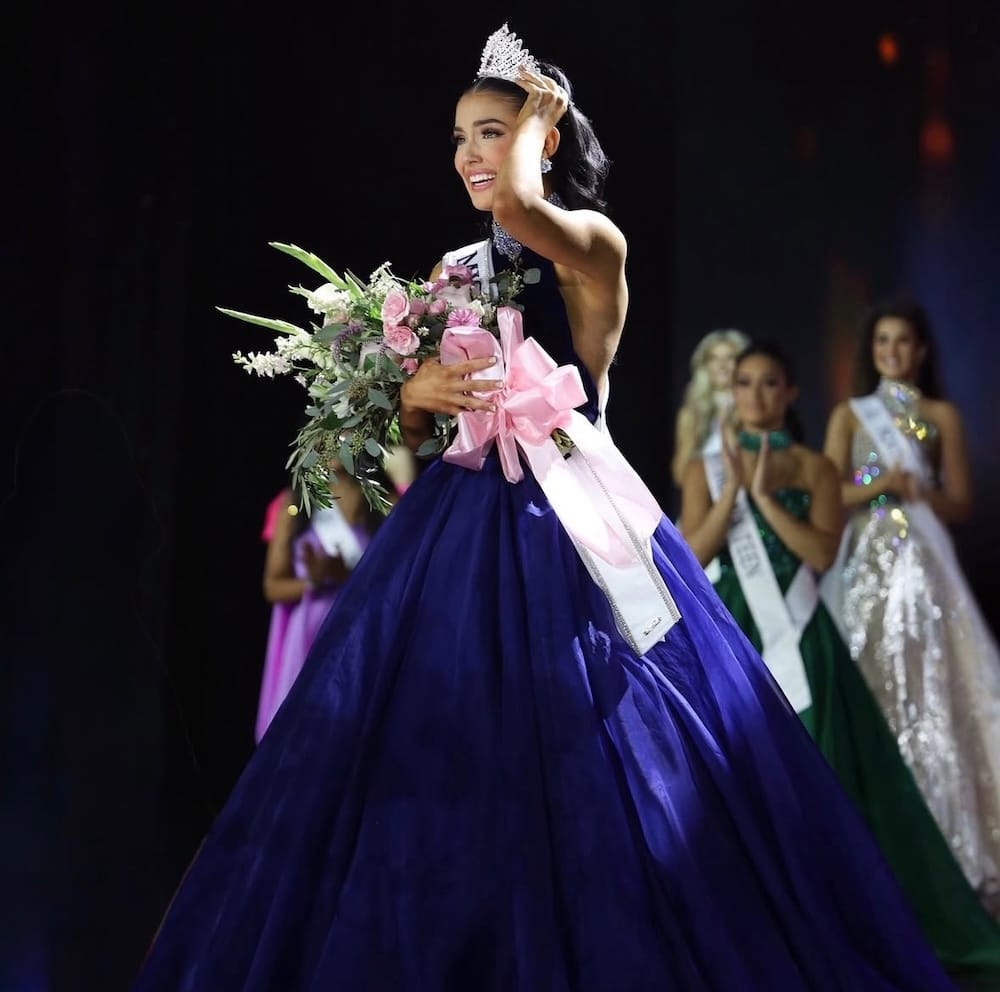 A woman in a blue ball gown and sash smiles, wearing a crown and holding a bouquet with pink ribbons on a stage, reminiscent of the crowning moment at Miss Teen USA. Other contestants in evening gowns stand blurred in the background.