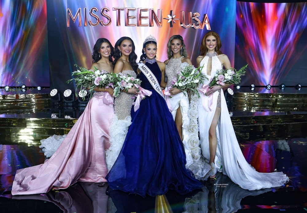 Five women in elegant gowns stand on stage holding bouquets, with one wearing a crown and a sash reading Miss Teen USA. The colorful backdrop displays MISS TEEN USA in large letters, capturing the excitement of the prestigious Miss Teen USA pageant.