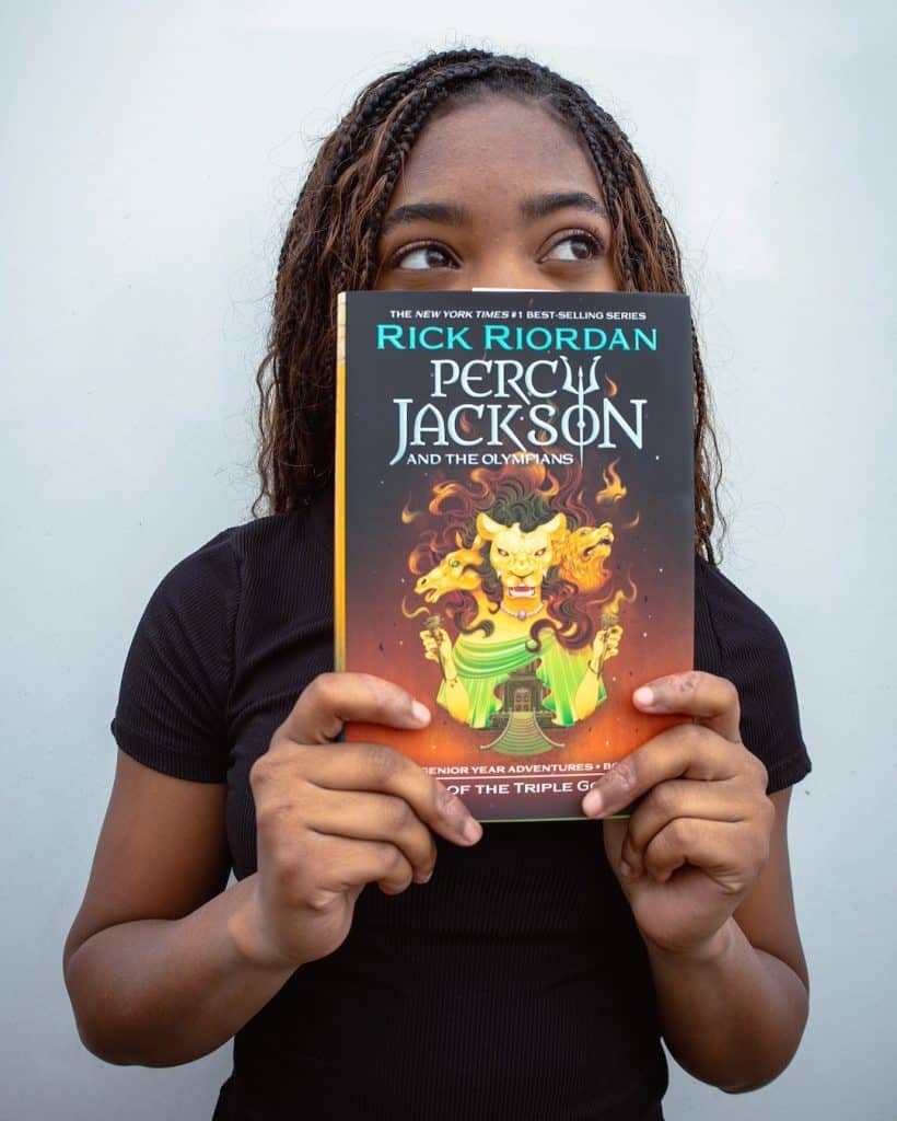A person with braided hair holds a Percy Jackson and the Olympians book, partially covering their face, against a plain white background.
